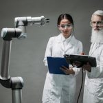Dois pesquisadores de jaleco analisam um braço robótico em um laboratório.