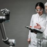 Pesquisadores usando jalecos e óculos de proteção analisam um braço robótico em um laboratório.