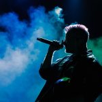Cantor em silhueta se apresenta no palco diante de luzes coloridas e fumaça cênica.