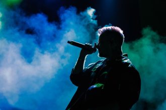 Cantor em silhueta se apresenta no palco diante de luzes coloridas e fumaça cênica.