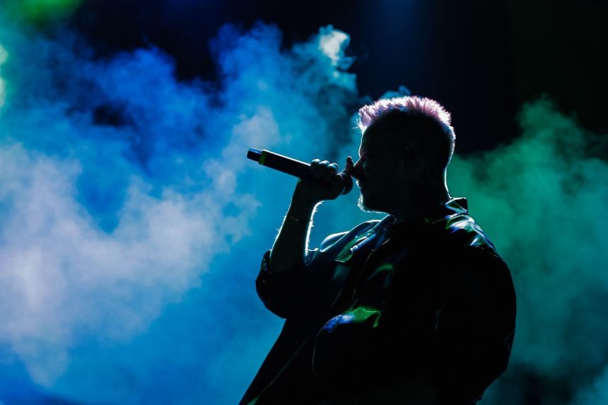 Cantor em silhueta se apresenta no palco diante de luzes coloridas e fumaça cênica.